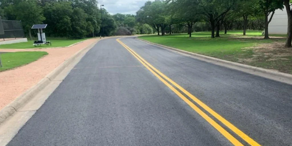 A newly paved road, crafted by expert asphalt contractors from Austin, features double yellow lines as it winds through a park area. Flanked by green grass and trees, a small solar-powered sign is visible on the left side.
