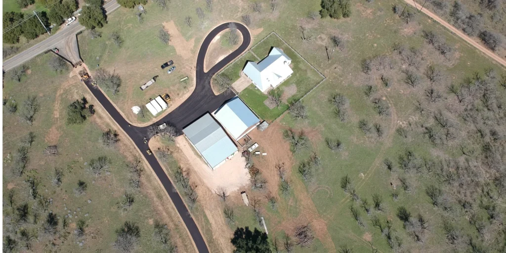 Aerial view of a rural property featuring a house, two metal buildings, and an asphalt driveway gracefully winding through the landscape. Scattered trees frame the scene, with a nearby road enhancing accessibility.