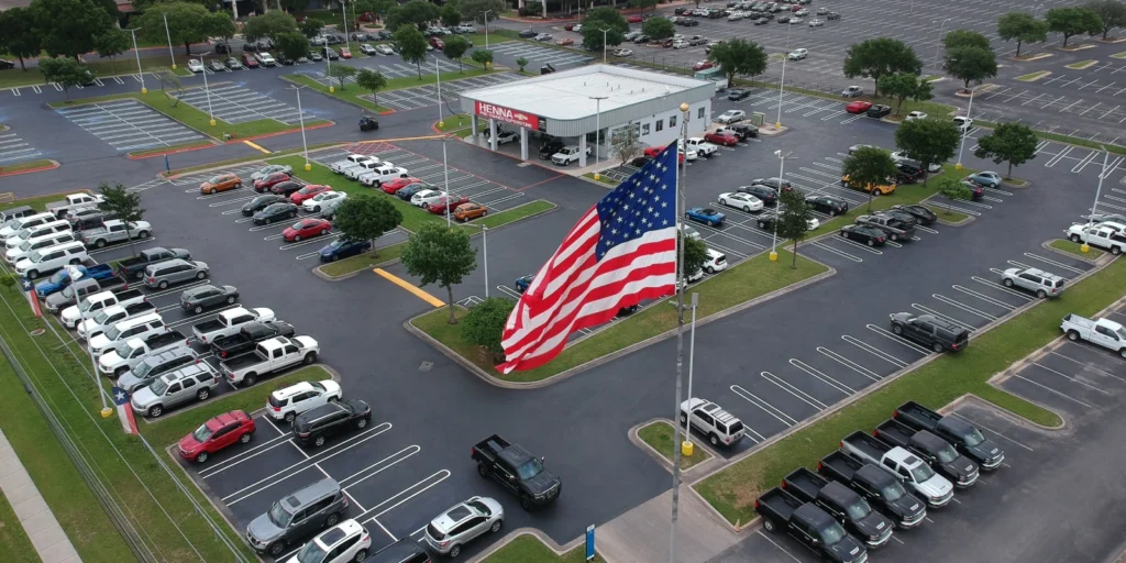 Aerial view of a parking lot with an American flag prominently displayed amid recent asphalt parking lot repairs. Several cars are parked near a gas station in the background.
