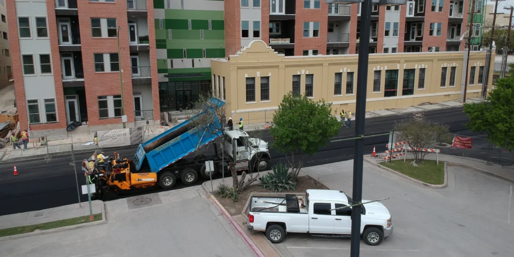 Construction workers in Austin are busy with asphalt paving, as a dump truck unloads into the paving machine. Nearby, several contractors' asphalt crew members ensure the street looks perfect in front of multistory buildings.