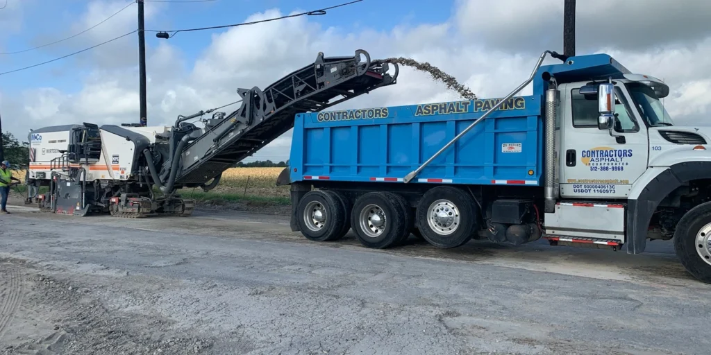 A machine from the Concrete Company loads asphalt into a blue dump truck on a roadwork site under a cloudy sky.