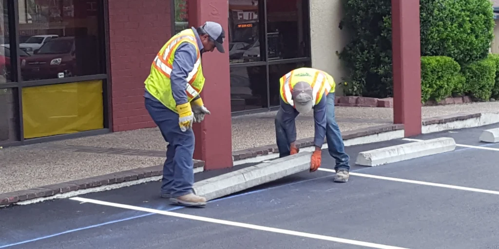 Two workers in safety vests install concrete wheel stops in a parking lot next to a building.
