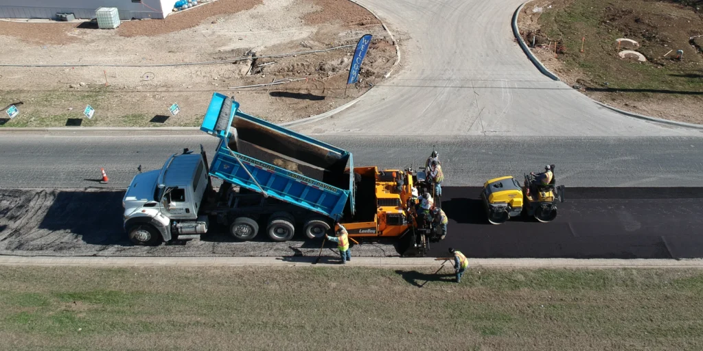 A dump truck pours asphalt for a crucial repair job, as construction workers and machinery expertly spread and compact the material.